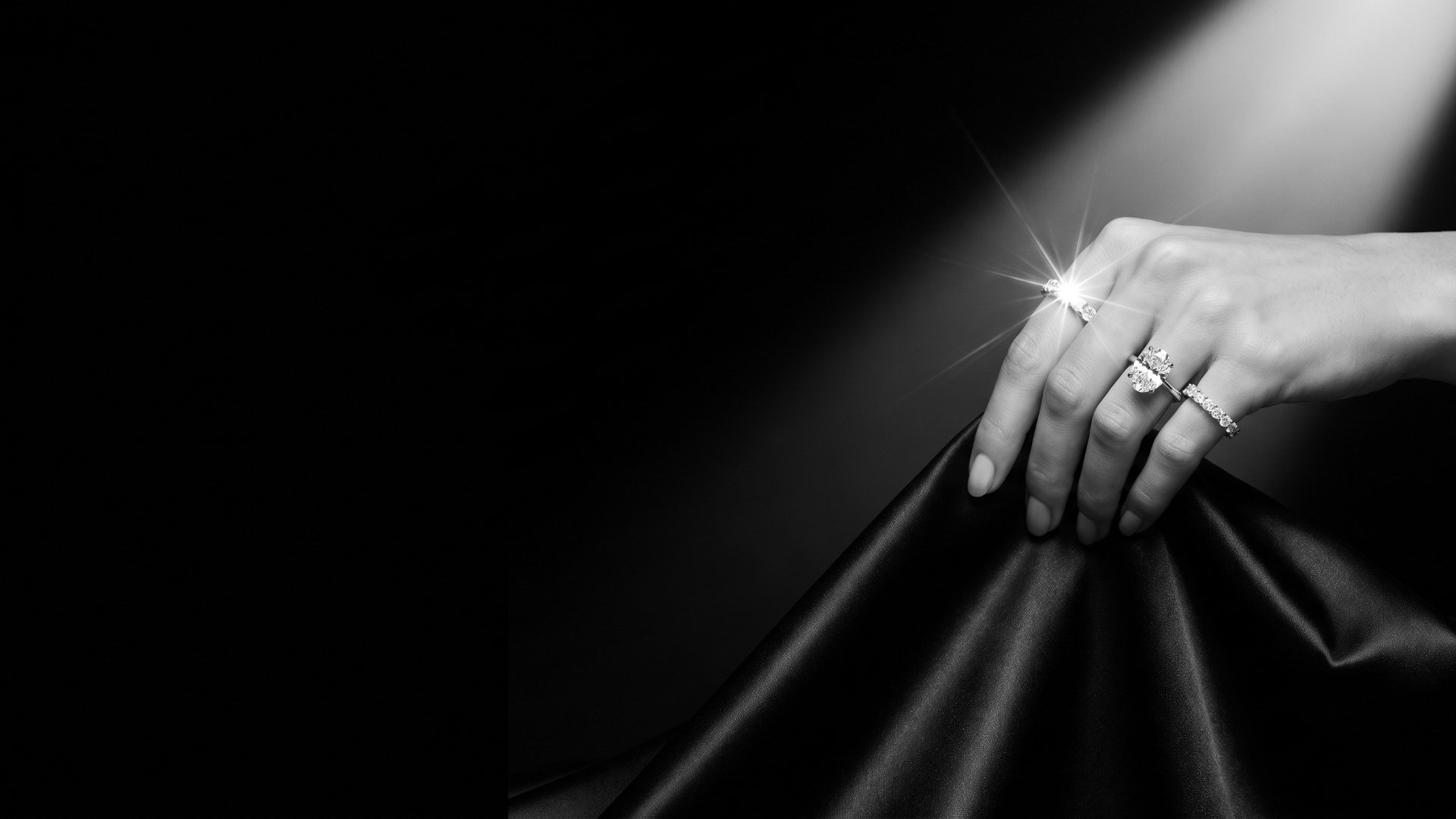 Hand with an oval diamond solitaire and eternity band lifting black silk under a spotlight