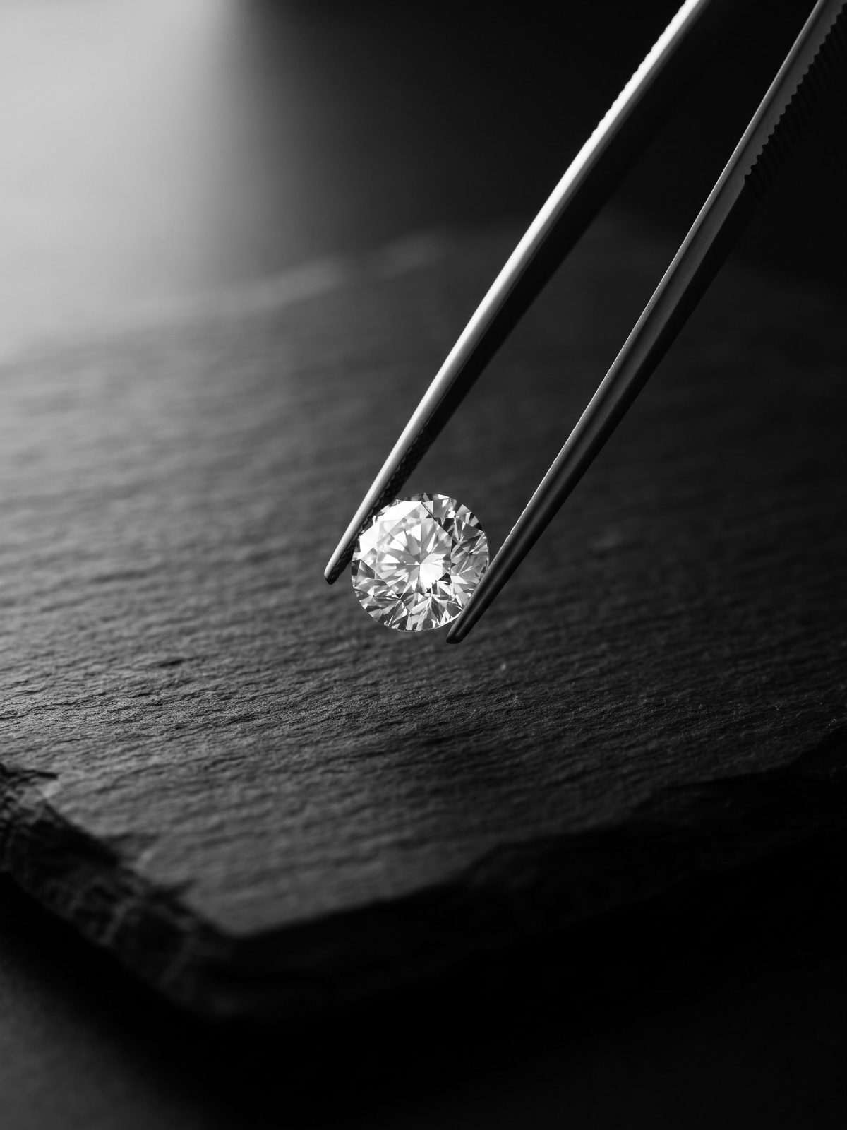 Loose round brilliant diamond held in steel tweezers above grey stone