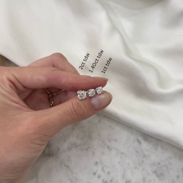 A hand holding three round diamond earrings, including a pair of 2ct tdw Round Brilliant Cut Lab Diamond Studs set in 14k White Gold, labeled with their carat sizes: 2 ct, 1.40 ct, and 1 ct. The diamonds rest on a white fabric background.