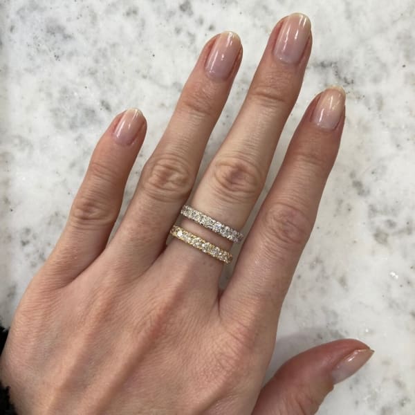 A hand with a light skin tone rests on a marble surface, adorned with two stacked rings. The first is the elegant 1.10ct Round Brilliant Cut Lab Diamond Wedding Ring in gold, and the other is a silver ring, both intricately designed with small embedded diamonds. The nails are neatly manicured with a natural finish.