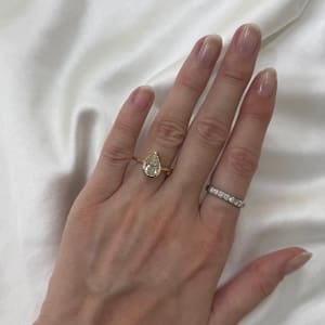 A hand adorned with a 1.59ct Pear Lab Grown Diamond Bezel Engagement Ring on one finger, accompanied by a thin, diamond-studded band on another, gracefully rests on white silk fabric.