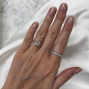 A hand showcases two rings: a 1.71ct Oval Lab Grown Diamond Engagement Ring on the ring finger and a diamond-studded band on the middle finger, set against a backdrop of white satin fabric and marble.