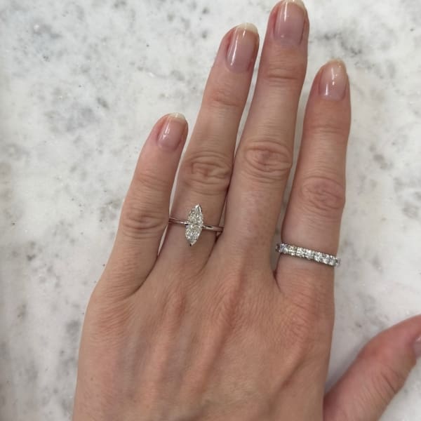 A hand showcases the stunning 1ct Marquise Solitaire Natural Diamond Engagement Ring on the ring finger, accompanied by a silver band with small diamonds on the middle finger. The hand is elegantly positioned on a light marble surface, with nails that are manicured and natural.