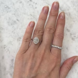 A hand with a 1ct Oval Natural Diamond Halo Engagement Ring on the ring finger and a diamond band on the index finger, placed on a white marble surface.