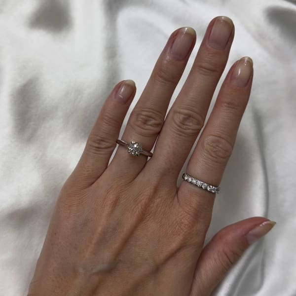 A hand with manicured nails rests on a white satin fabric. The index finger displays the 1ct Round Brilliant Cut Natural Diamond Engagement Ring, while the ring finger features a band with small diamonds. The nails are natural and slightly pointed.