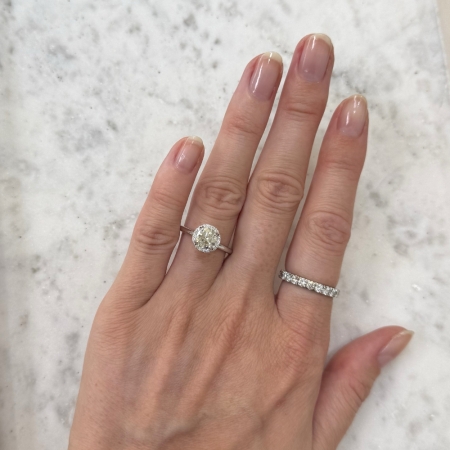 A hand adorned with a 1ct Round Brilliant Cut Natural Diamond Halo Engagement Ring on the ring finger and a diamond-encrusted band on the middle finger is elegantly placed on a light marble surface. The nails are manicured, showcasing a natural style.