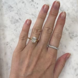 A hand displayed over a marble surface, showcasing two rings: the "2.05ct Oval Lab Grown Diamond Engagement Ring With Hidden Halo," a gold band with a prominent oval diamond on the ring finger, and a silver band adorned with small stones on the middle finger. The nails are manicured and natural.