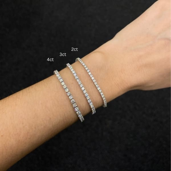 A wrist adorned with three diamond tennis bracelets of varying sizes, including a 2.14ct Lab Diamond Tennis Bracelet in 14k White Gold, against a black background.