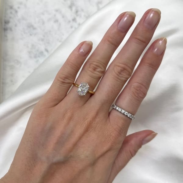 A hand showcasing two rings: on the ring finger, there is a gold band adorned with a 2.27ct Oval Lab Diamond, and on the middle finger, a silver band embellished with smaller diamonds. The backdrop is a soft white fabric.