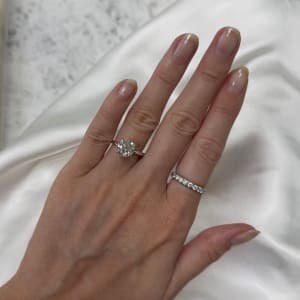 A hand with beautifully manicured nails showcasing two rings. The first ring is the 2.50ct Round Lab Diamond Engagement Ring Hidden Halo on the ring finger, and the second is a band adorned with multiple small diamonds on the middle finger. The background is a soft, white fabric.