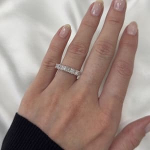 A hand with neatly manicured nails is shown wearing the 2.52ct Cushion Lab Diamond Half Eternity Ring, featuring a row of sparkling diamonds set in silver. The background displays soft, white fabric, and the person is adorned in a black long-sleeve garment.