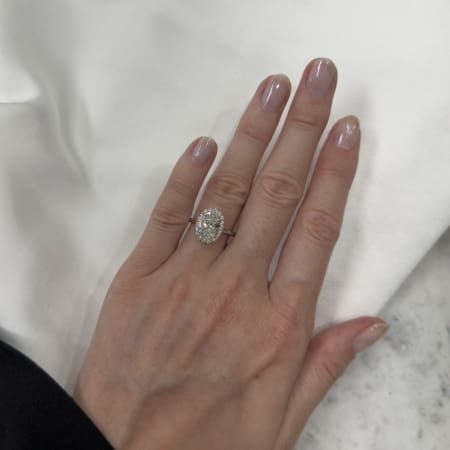 A hand with neatly manicured nails rests on a white fabric, adorned with an elegant 2.05ct oval lab grown diamond engagement ring featuring a 0.28ct halo on the ring finger, set in a simple band.