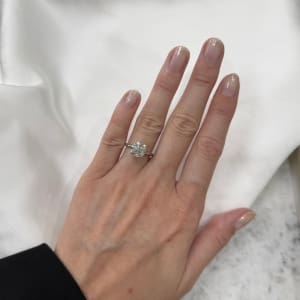 A hand with neatly manicured nails is wearing a large, sparkling diamond ring on the ring finger. The background is a soft white fabric.