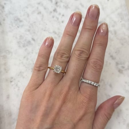 A hand with neatly manicured nails wearing two rings on the ring finger: a gold band featuring a stunning 2ct cushion natural diamond solitaire with a hidden halo and a silver band adorned with multiple smaller diamonds. The background is a light, speckled surface.