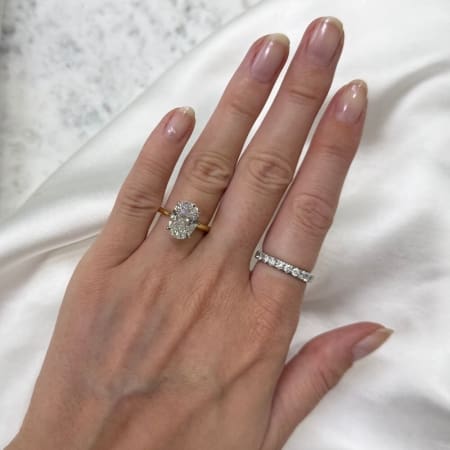 A hand showcasing two rings on the ring finger: one featuring a 3.53ct oval-shaped diamond perched on a gold band, known as the Oval Lab Diamond Engagement Ring with Hidden Halo, and the other a silver band embellished with smaller diamonds. The backdrop consists of white fabric intermingled with a marble-like surface.