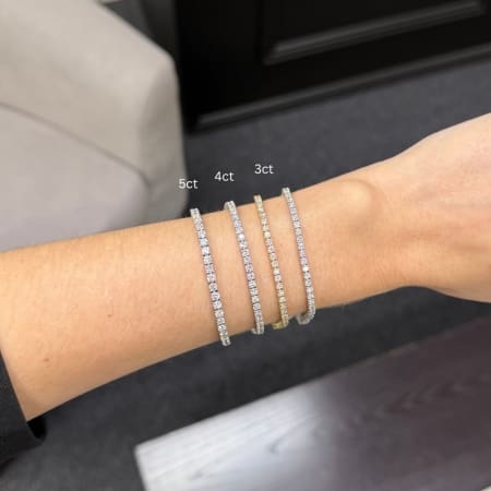 A hand wears a tennis bracelet labeled as a 4ct Lab Grown Diamond Tennis Bracelet in 14k White Gold, among others labeled as 5ct and 3ct. Each bracelet is adorned with sparkling stones, decreasing in size from left to right. The background features neutral-colored furnishings.