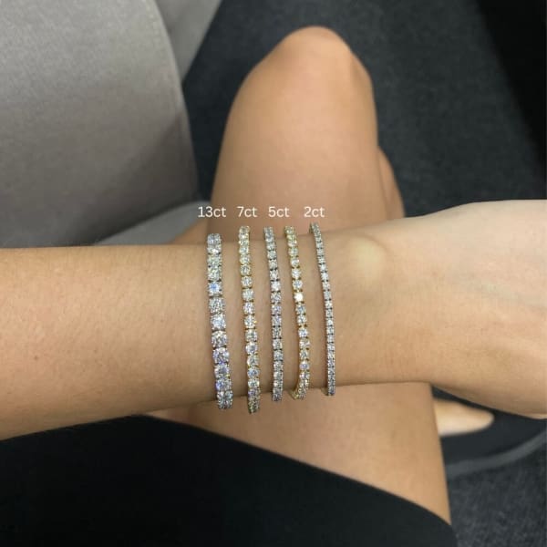 A person's arm is adorned with five tennis bracelets featuring lab-grown diamonds, each labeled with its carat weight: 13ct, 7ct, the 5.27ct bracelet in 14k yellow gold, and 2ct, from top to bottom. The person's crossed legs are visible in the background.