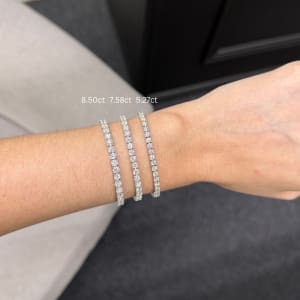 A hand is adorned with three diamond tennis bracelets of varying sizes, beautifully showcasing their carat weights: 8.50 ct, 7.58 ct, and 5.27 ct. Above one of the bracelets, you notice the inscription "7.85ct Lab Grown Diamond Tennis Bracelet in 14k White Gold." The background features a soft chair against a dark wall backdrop.