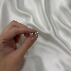 A hand holding two large, dazzling Certified 3ct tdw Round Brilliant Cut Lab Diamond Studs against a backdrop of white satin fabric.