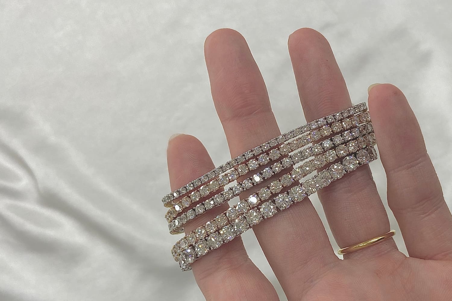 A hand with a gold ring holds three sparkling diamond bracelets against a white, satin-like background.