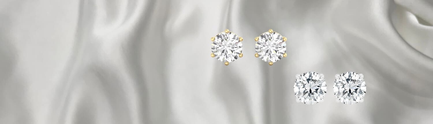 Four diamond earrings are displayed against a smooth, gray satin background. Two earrings have gold prongs, while the other two have a simpler setting, showcasing their multifaceted brilliance.