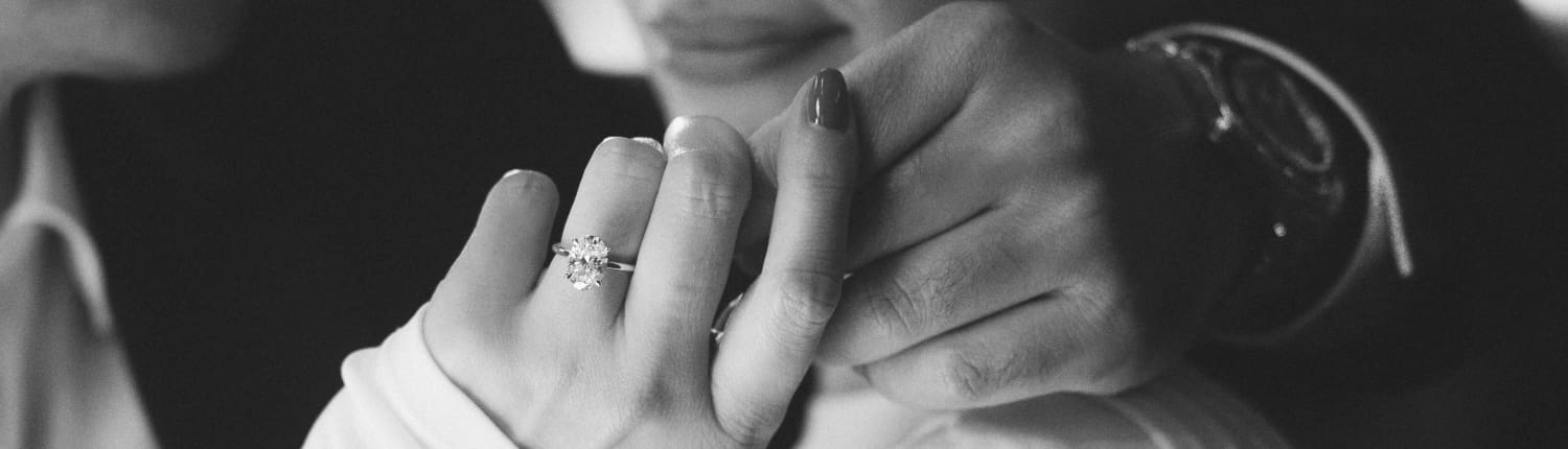 A close-up black and white photo of two people gently holding hands. One hand features a large, sparkling engagement ring. The scene conveys a sense of intimacy and connection.