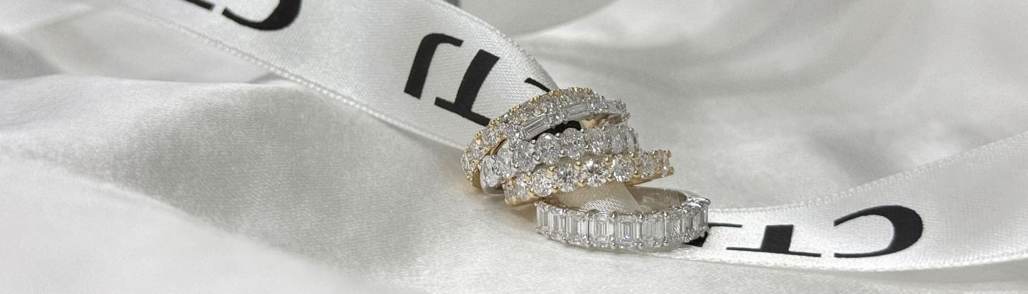 A collection of sparkling diamond rings in various styles sits on a white satin ribbon. The rings feature different cuts and settings, including round and emerald cut diamonds, against a soft, elegant background.